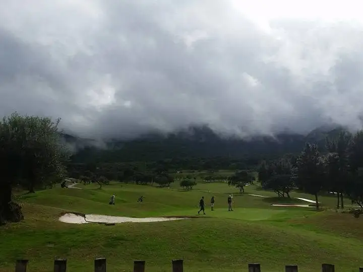 Golfkenttä oliivipuiden välistä nähtynä, matalat pilvet peittävät vuorten huiput ja pelaajat kävelevät väylällä