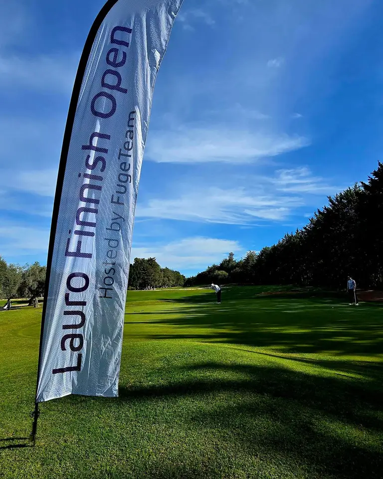 Lauro Finnish Open hosted by FugeTeam -banderolli väylän reunassa aamuauringossa, taustalla pelaajia