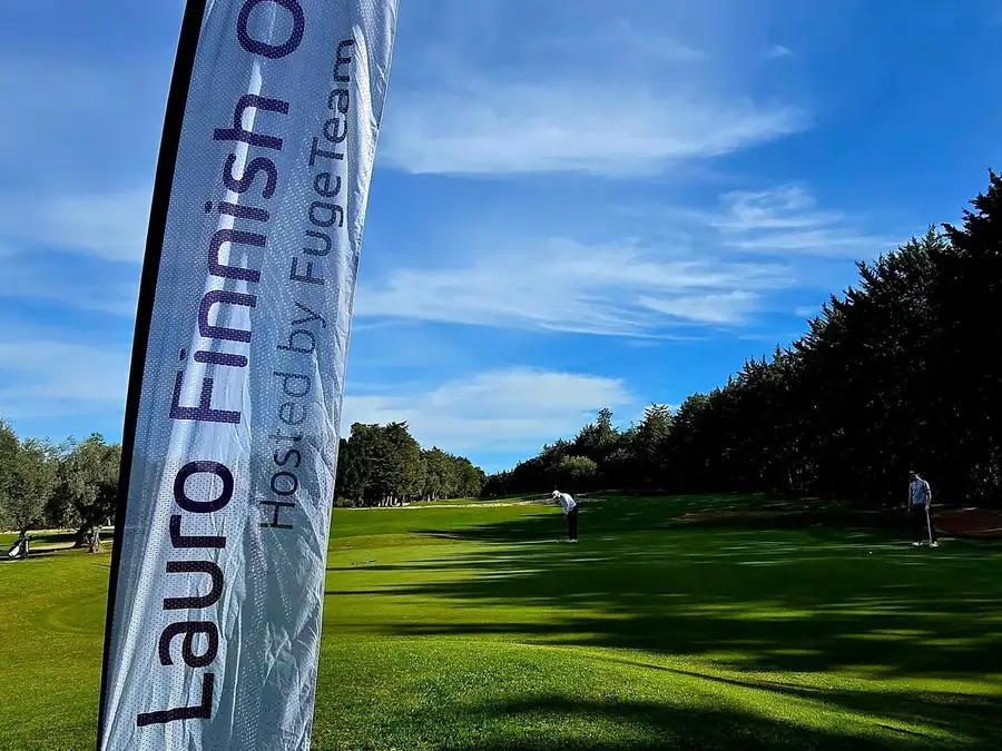 Lauro Finnish Open hosted by FugeTeam -banderolli väylän reunassa aamuauringossa, taustalla pelaajia