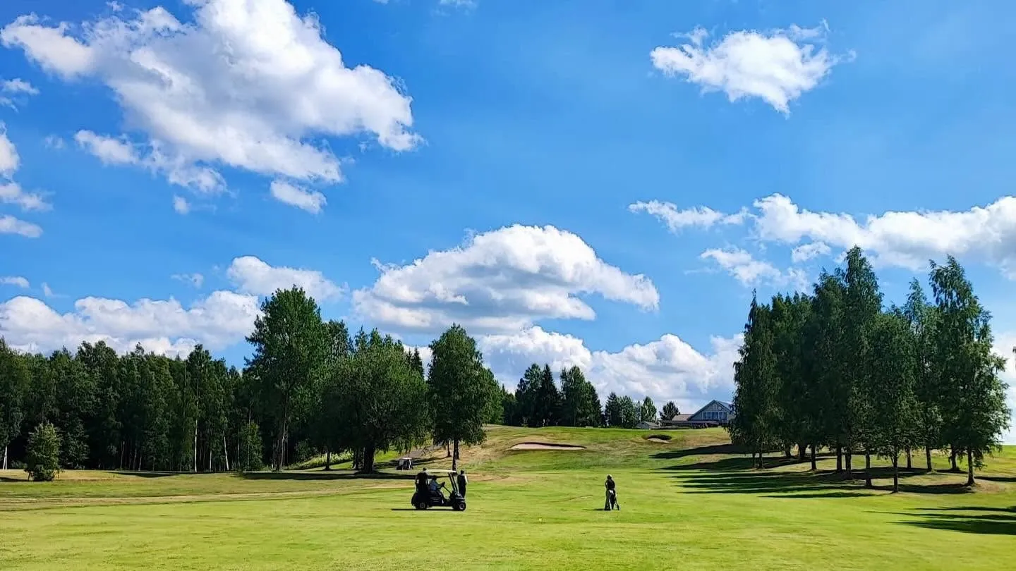 Vihreä väylä kesäpäivänä, taustalla kuusia, golfauto ja poutapilviä sinisellä taivaalla