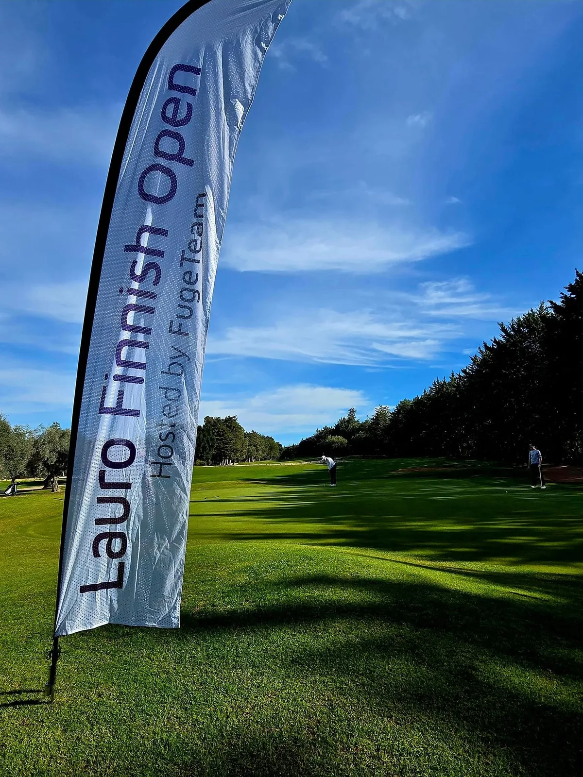 Lauro Finnish Open hosted by FugeTeam -banderolli väylän reunassa aamuauringossa, taustalla pelaajia